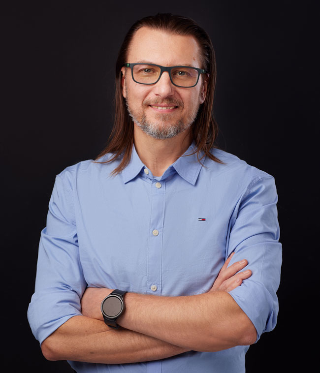 Mężczyzna w okularach, w niebieskiej koszuli z złożonymi rękami, stoi na ciemnym tle, uśmiechnięty.