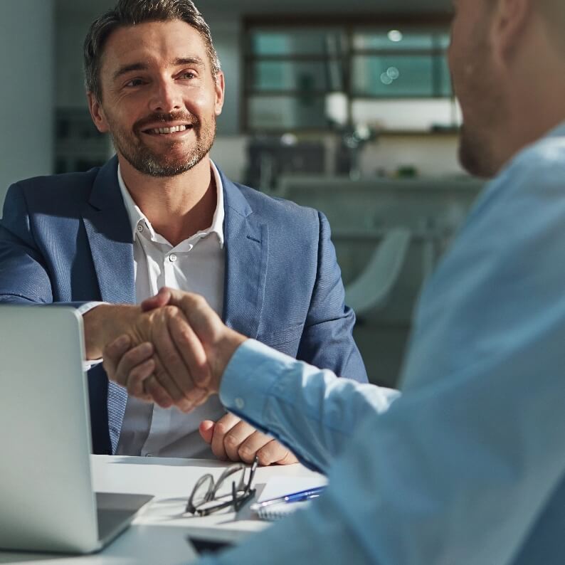 Dwóch mężczyzn w biurze podaje sobie ręce, uśmiechając się przy komputerze. Na stole okulary i długopis.