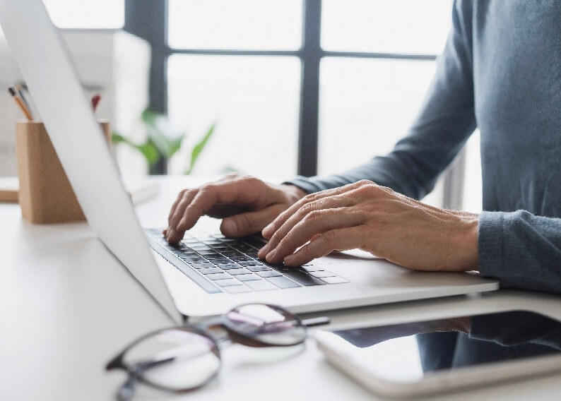 Osoba pisząca na laptopie, okulary leżą obok na biurku. W tle widać rozmytą roślinę i okno.