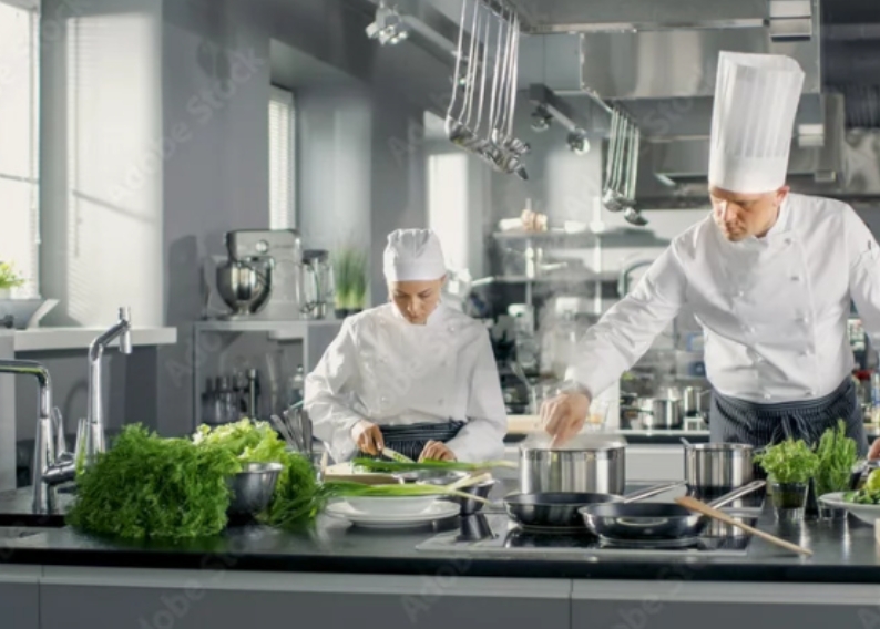 Szefowie kuchni w jasnej kuchni. Jedna osoba kroi warzywa, druga gotuje przy garnkach. Wokół świeże zioła.